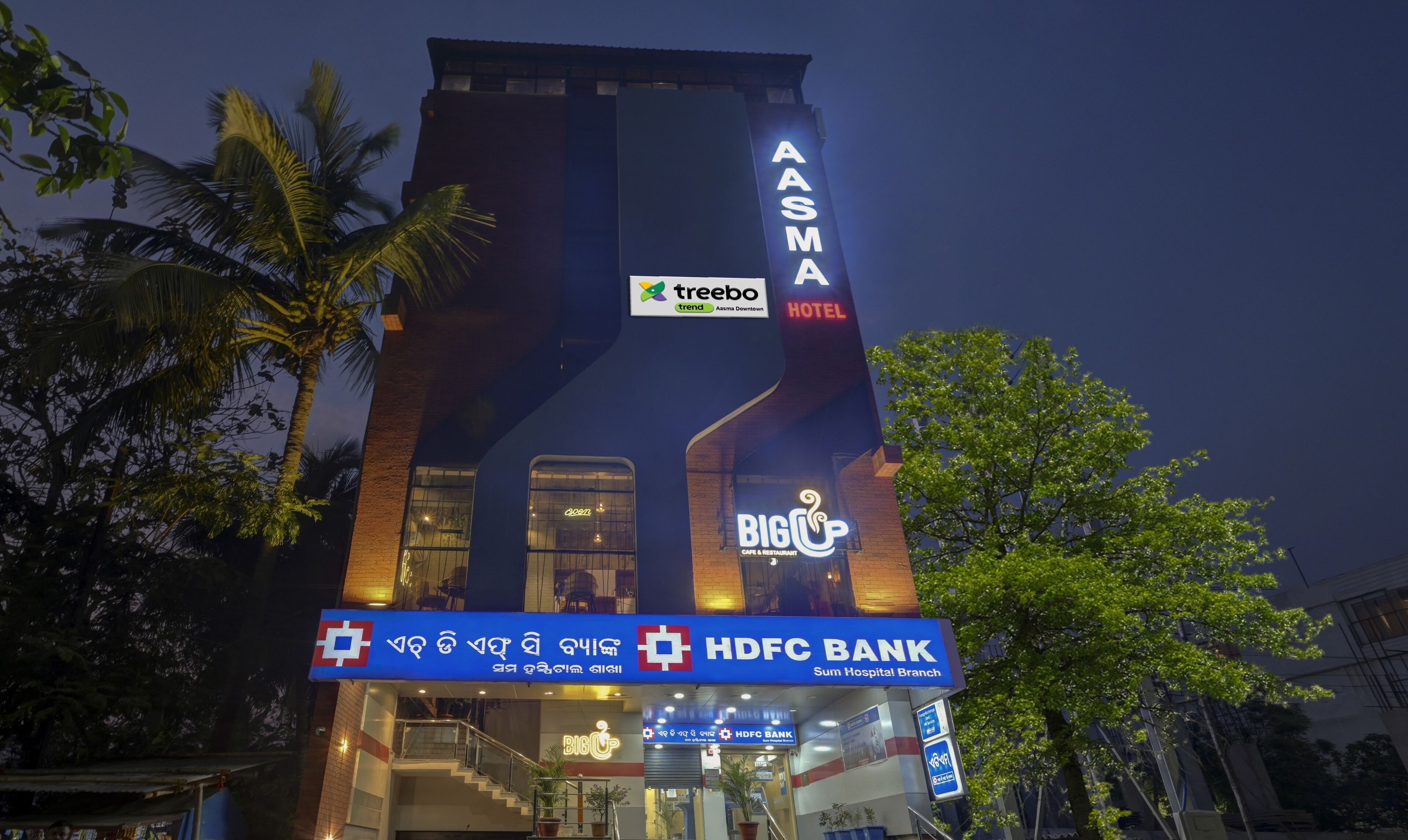 A building with a sign for AASMA HOTEL, a TREEBO restaurant, and HDFC BANK at the Sum Hospital branch.