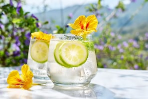 To glas med en drik, limeskiver og blomster, placeret på et bord med en uskarpt naturlandskab i baggrunden.