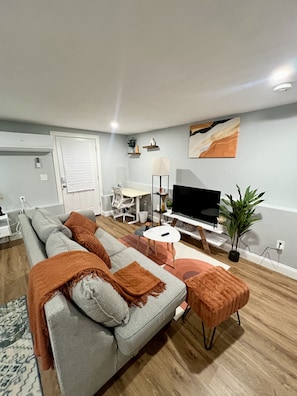 A modern living room with a grey sofa, orange throw pillows, a small white table, a TV, and a plant.