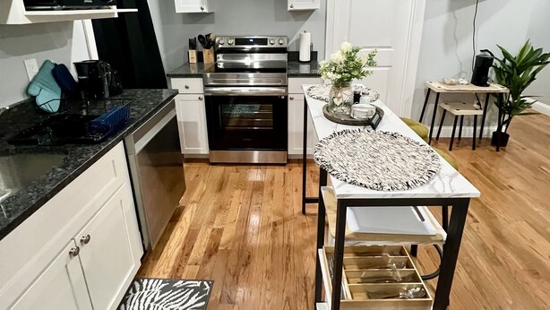 Una cuina moderna amb armaris blancs, electrodomèstics d'acer inoxidable, un terra de fusta i una zona de dinar amb taula i caires.