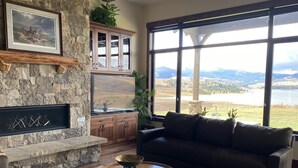 A living room with a stone fireplace, a large window with a view of a lake and mountains, a dark leather sofa, and a wooden coffee table. - Views, views, views!!!  Secluded setting over-looking beautiful Steamboat Lake. (Clark)