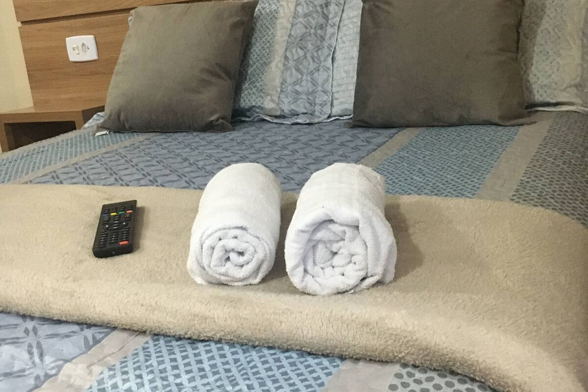 Cama bem arrumada com um cobertor, duas toalhas enroladas e um controle remoto.