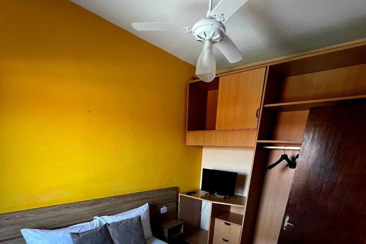 Uma cama bem arrumada com roupa de cama e toalhas brancas, um ventilador de teto, uma televisão sobre um suporte de madeira e um guarda-roupa.