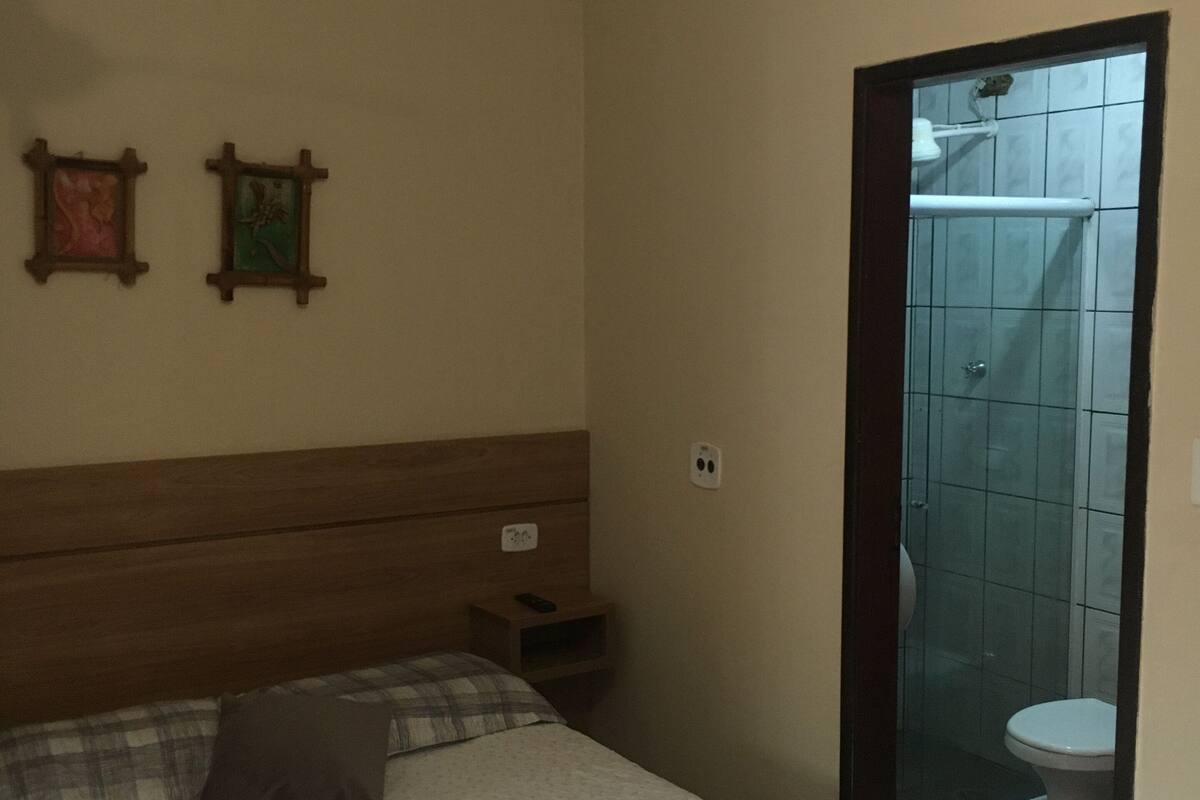 Quarto com cama, ventilador de teto, mesa de cabeceira e banheiro visível através de uma porta aberta.