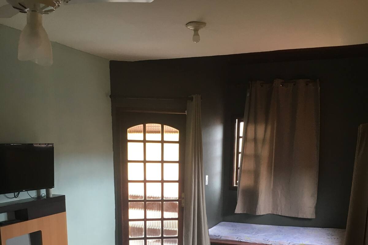 Quarto com cama, televisão em suporte, janela com cortinas e ventilador de teto.
