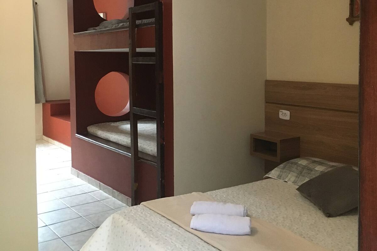 Quarto de hotel com cama, ventilador de teto, cabeceira de madeira, mesa de cabeceira e prateleira embutida com recortes circulares.