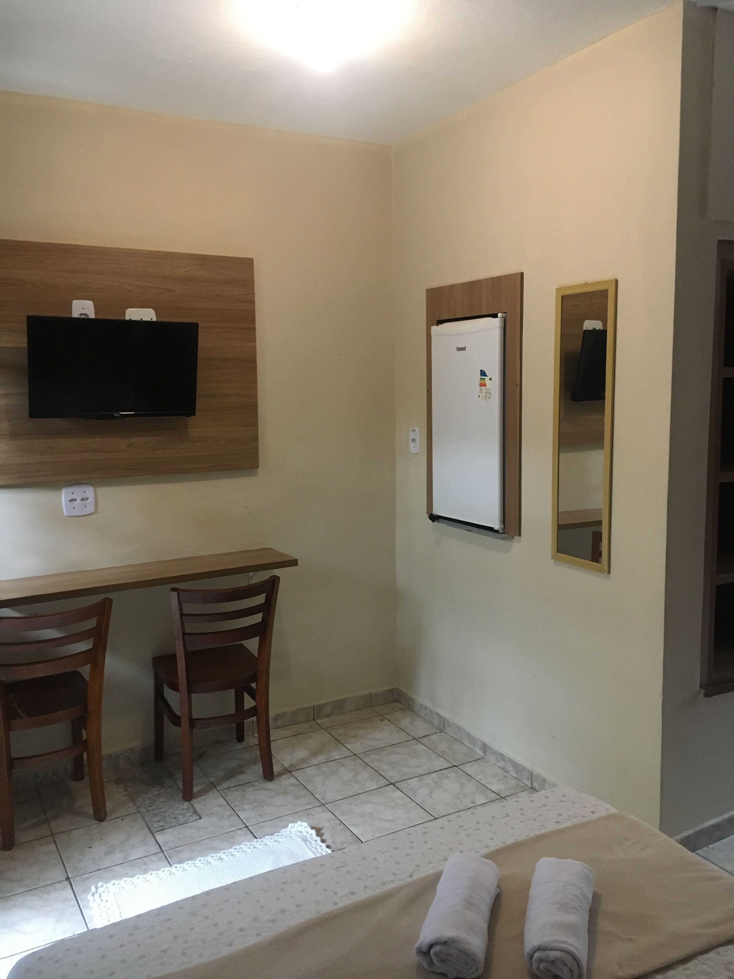 A hotel room with a bed, two wooden chairs, a desk, a television mounted on the wall, a refrigerator, and a mirror.