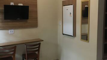 A hotel room with a bed, two wooden chairs, a desk, a television mounted on the wall, a refrigerator, and a mirror.