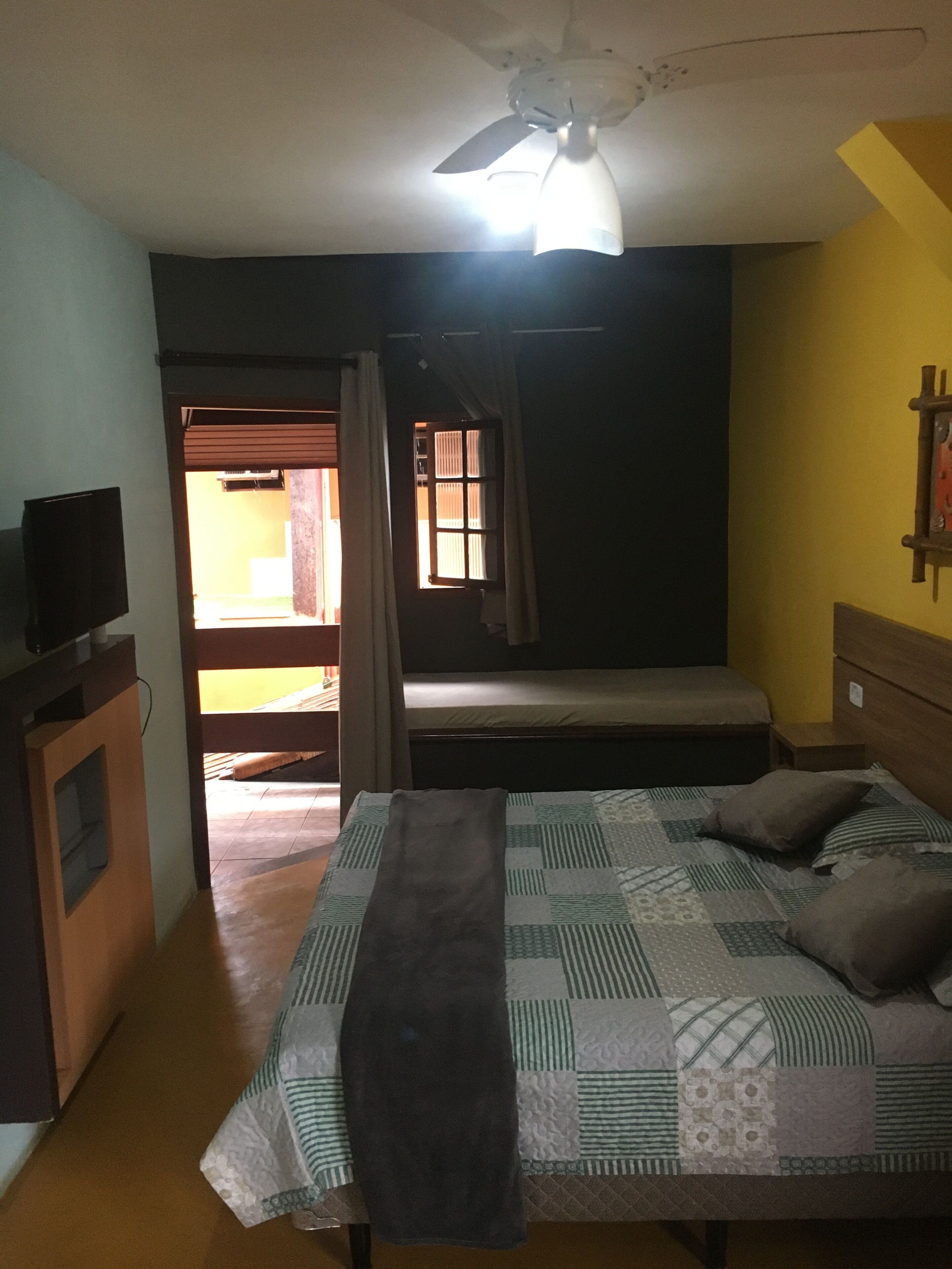 Un dormitorio con cama, televisor, ventilador de techo y ventana con cortinas.