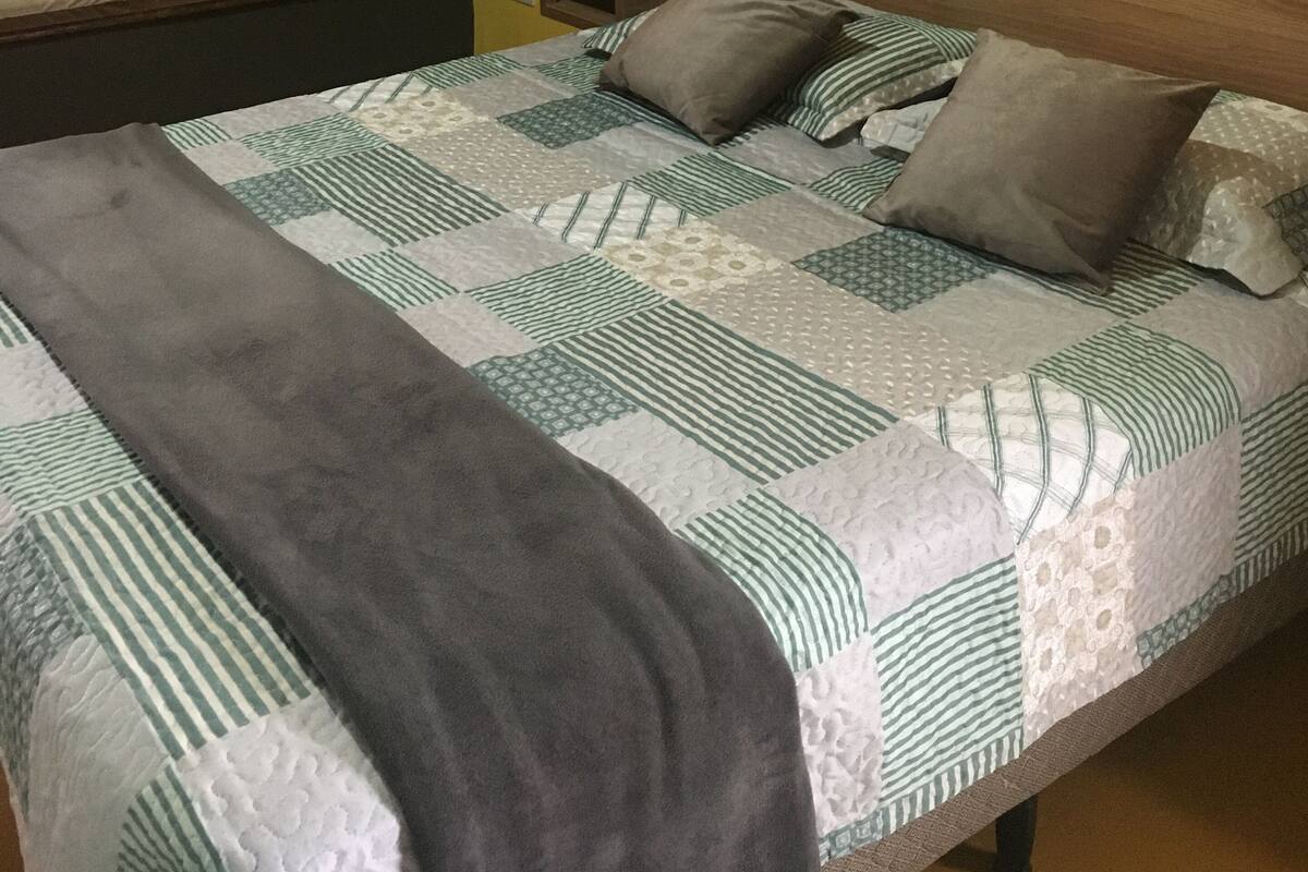 Cama com colcha, travesseiros e cobertor. Tem uma mesinha de cabeceira com controle remoto.