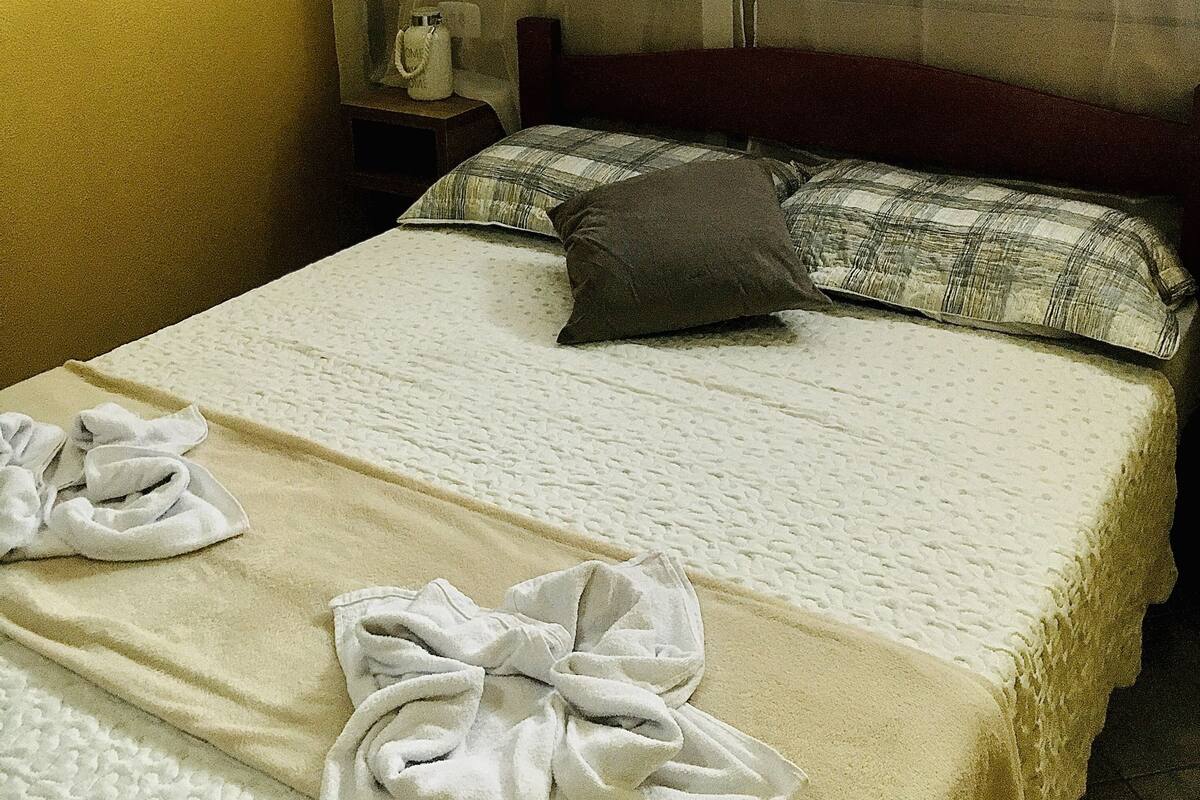 Quarto de hotel com cama, mesa de cabeceira e janela com cortinas translúcidas.