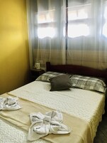 Habitación de hotel con cama, mesita de noche y ventana con cortinas traslúcidas.