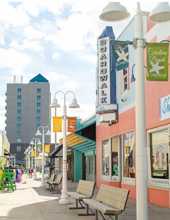 A boardwalk with shops and benches, a tall building in the background, and a clear sky.