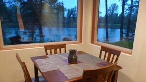 A dining table with a woven placemat, surrounded by wooden chairs, set against a window with a view of a forest and a body of water.