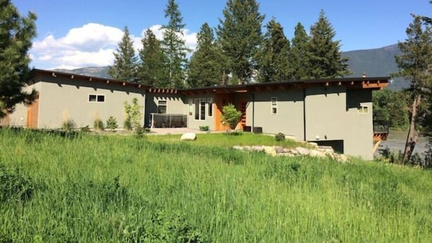 A modern two-story house with a covered patio, surrounded by tall trees and a grassy field.