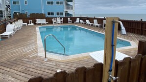 Un área de piscina con cubierta de madera, sillas reclinables blancas y un edificio azul al fondo.