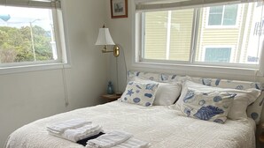 Una cama bien tendida con una colcha blanca y almohadas a juego, una lámpara de noche y un cuadro en la pared.