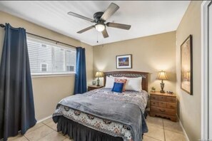 Un dormitorio con cama, ventilador de techo, ventana con cortinas, mesita de noche con lámpara y un cuadro enmarcado en la pared.