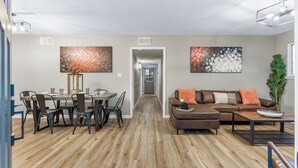 A modern living room with a brown leather sofa, a dining area with a round table and chairs, and two abstract paintings on the walls. - 3 miles from NAS * King and Queen Bed * Covered Patio * Luxury Bath * Open concept Living (Pensacola)
