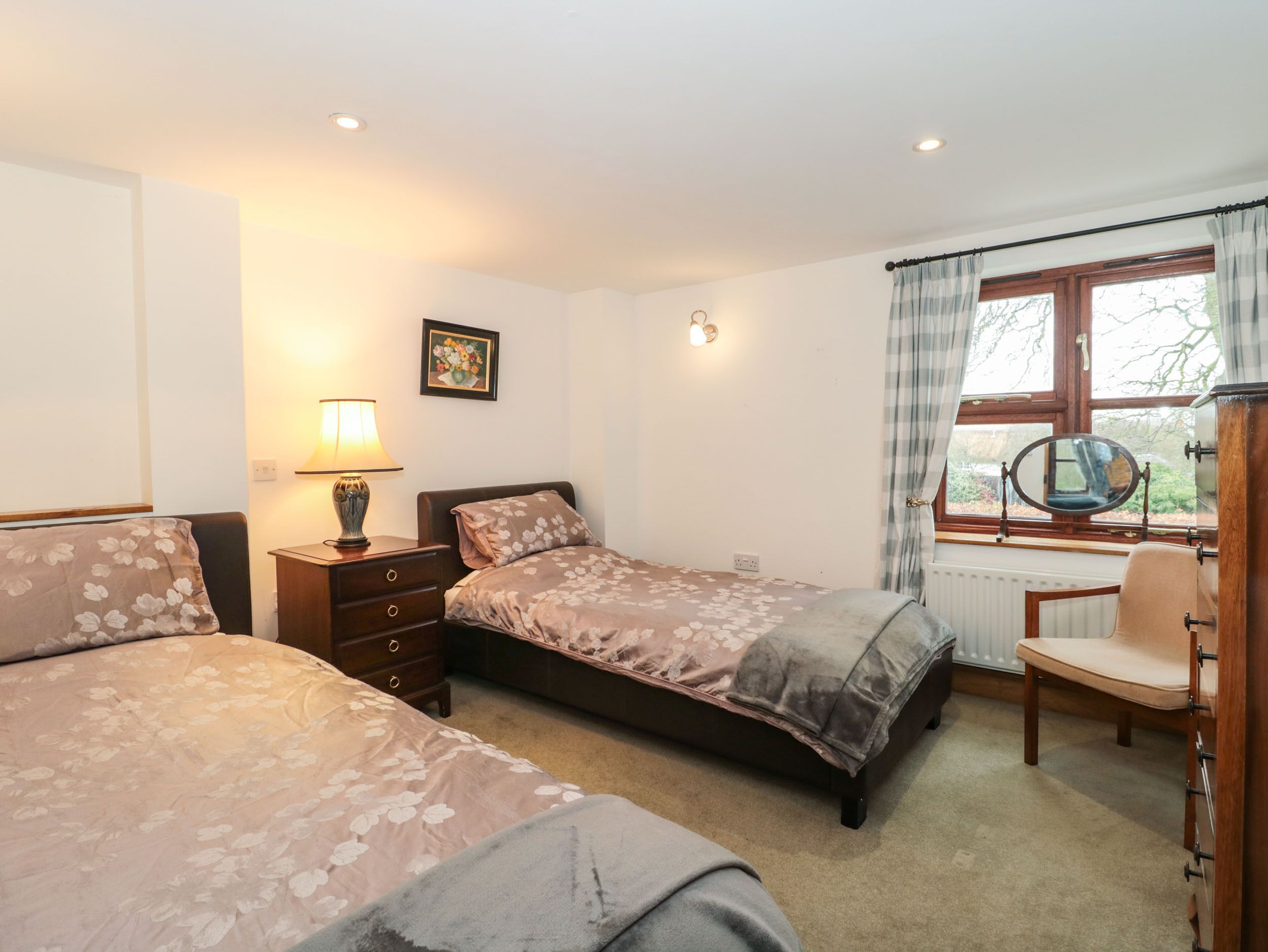 A bedroom with two beds, a desk with a chair, a window with curtains, a radiator, and a wall-mounted lamp.