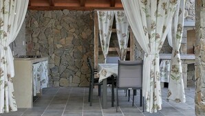 Un área de comedor al aire libre con techo, paredes de piedra, una mesa con sillas y cortinas florales.