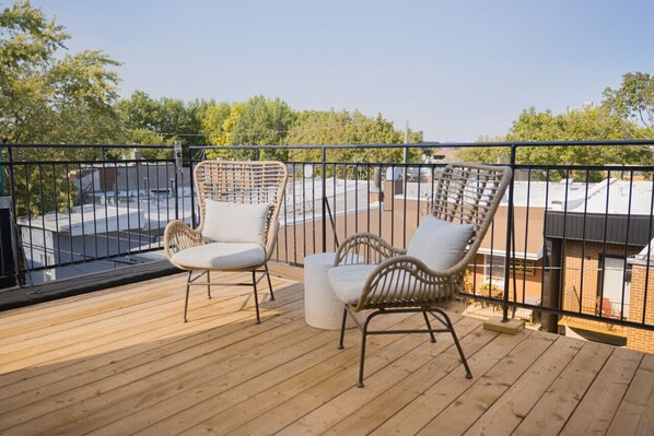 A wooden deck with two wicker chairs and a small table, overlooking a cityscape.
