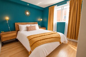 Une chambre avec un lit en bois, une literie blanche, un plaid jaune et un mur turquoise orné d’un miroir en forme de soleil.
