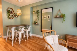 Un coin repas avec une table en bois et des chaises blanches, un fauteuil en osier, un luminaire suspendu et des étagères murales ornées de plantes.