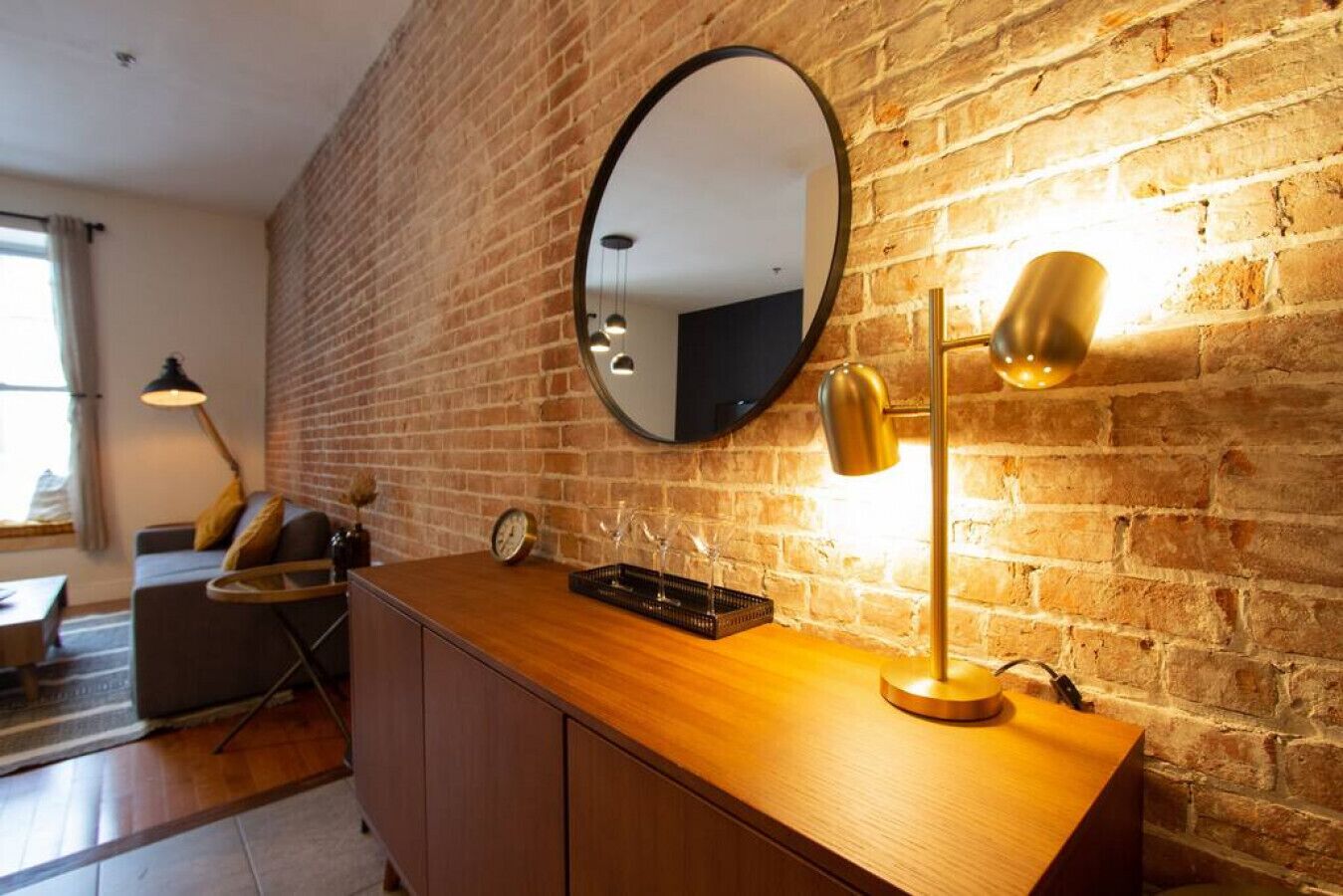Una habitación con pared de ladrillo, un espejo redondo, una consola de madera con una lámpara y un sofá con cojines.