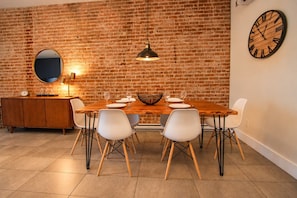 A dining area with a brick wall, a wooden table, white chairs, a round mirror, and a clock. - Vintage/Rooftop/Terraces/Plateau/St-Denis/AC/TV (Montréal)