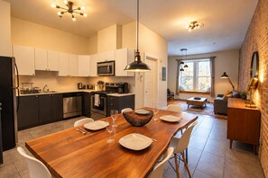 A modern kitchen with white cabinets, a wooden dining table set for four, and a brick wall. - Vintage/Rooftop/Terraces/Plateau/St-Denis/AC/TV (Montréal)