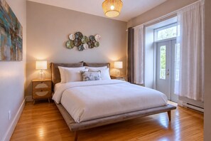A bedroom with a bed, two bedside tables, a wall art piece, and a large window with curtains. - Zen/Rooftop/Terraces/Plateau/St-Denis/AC/TV (Montréal)