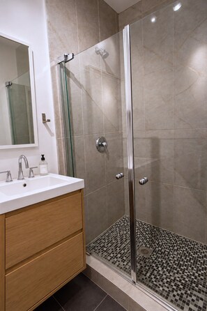 A bathroom with a glass shower enclosure, a white sink with a mirror above it, and a wooden vanity. - Zen/Rooftop/Terraces/Plateau/St-Denis/AC/TV (Montréal)