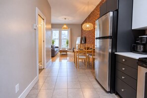 A modern kitchen with a stainless steel refrigerator, a coffee maker, and a dining area with wooden chairs and a table. - Zen/Rooftop/Terraces/Plateau/St-Denis/AC/TV (Montréal)