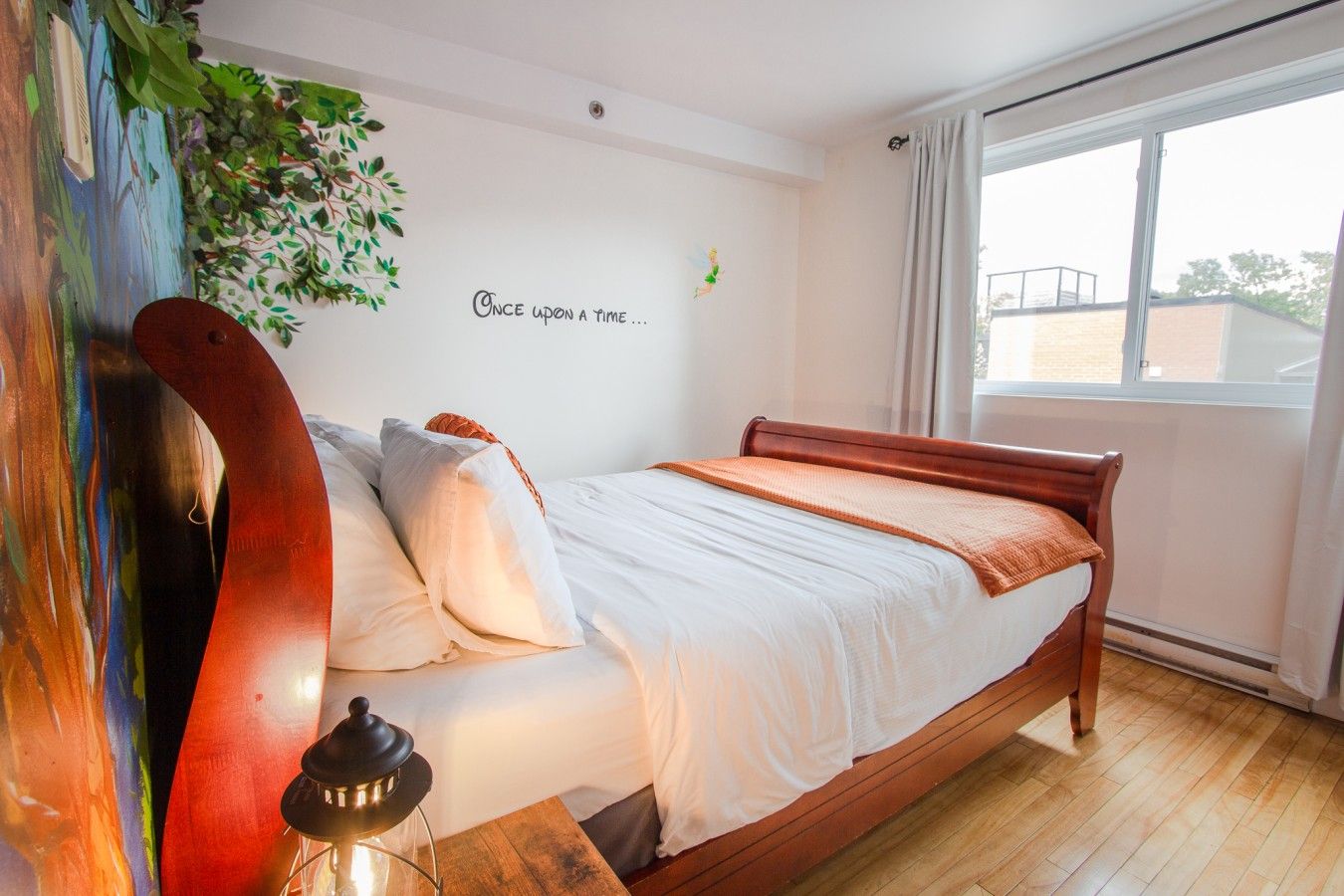 A bedroom with a bed, a wall mural, a window, and a view of the outside.
