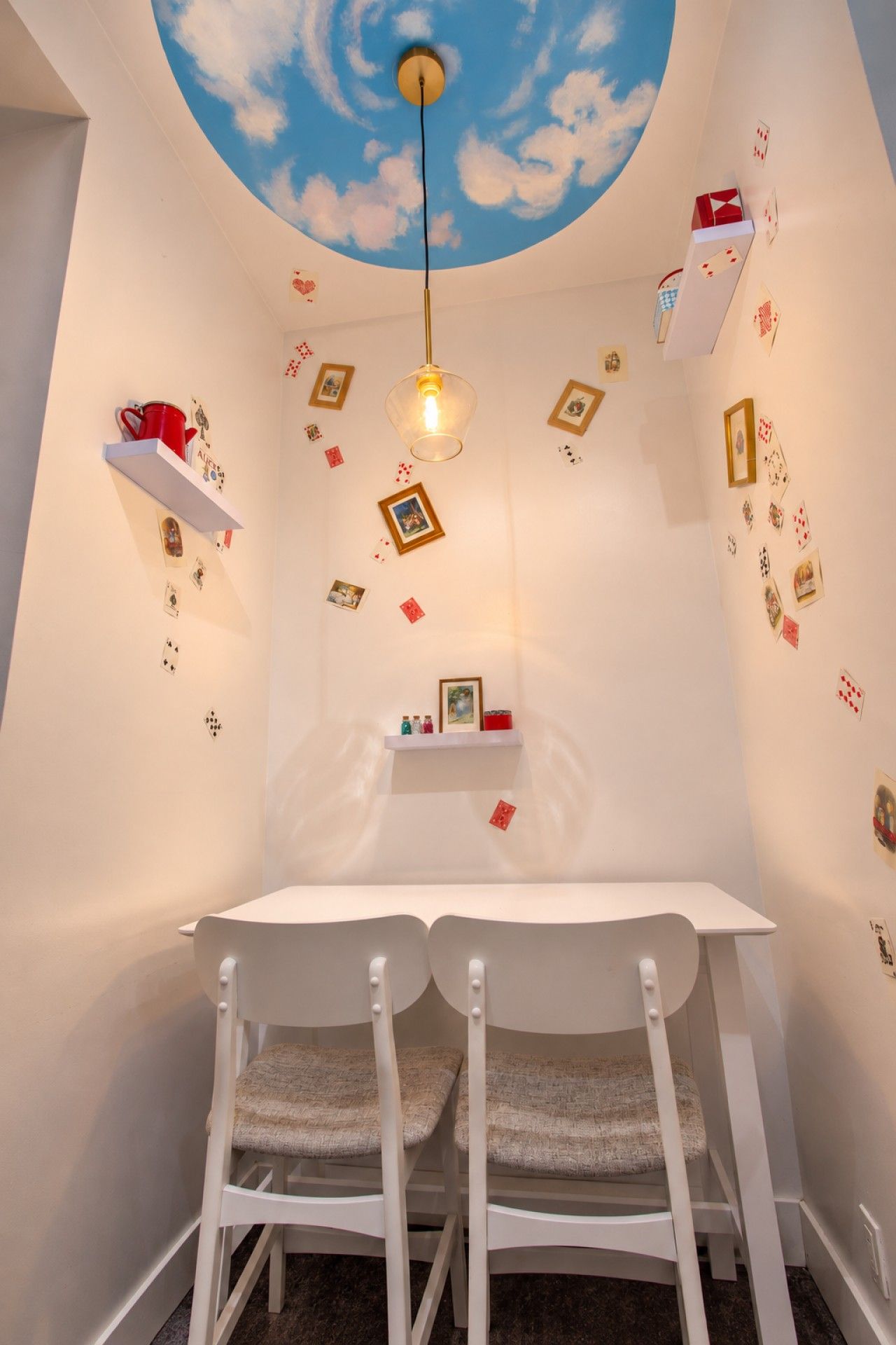 A room with a white table, two white chairs, a ceiling with a painted sky and clouds, and scattered playing cards on the walls.