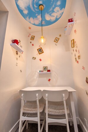Une pièce avec une table blanche, deux chaises blanches, un plafond orné d’un ciel peint et de nuages, et des cartes à jouer éparpillées sur les murs.