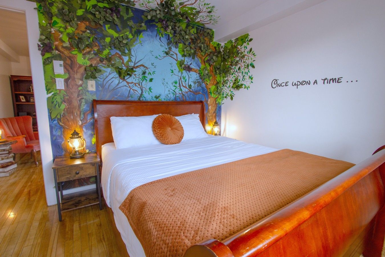 Een slaapkamer met een bed, een nachtkastje, een rode stoel, een boekenkast en een muur met een boomdecoratie.