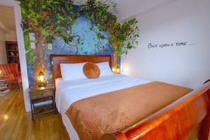 Une chambre à coucher avec un lit, une table de chevet, un fauteuil rouge, une bibliothèque et un mur orné d’une fresque représentant un arbre.