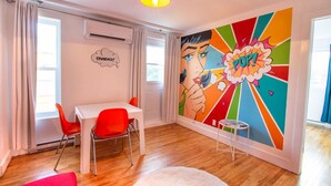 A modern room with a white table and red chairs, a colorful pop art mural, and a ceiling light. - Pop/Rooftop/Terraces/Plateau/St-Denis/AC/TV (Montréal)