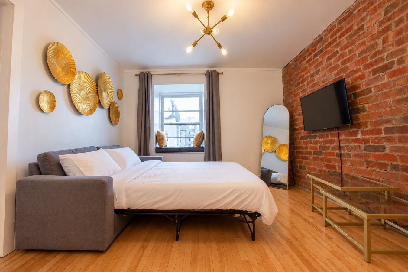 A modern bedroom with a bed, a grey sofa, a flat-screen TV, a brick wall, and a large window.