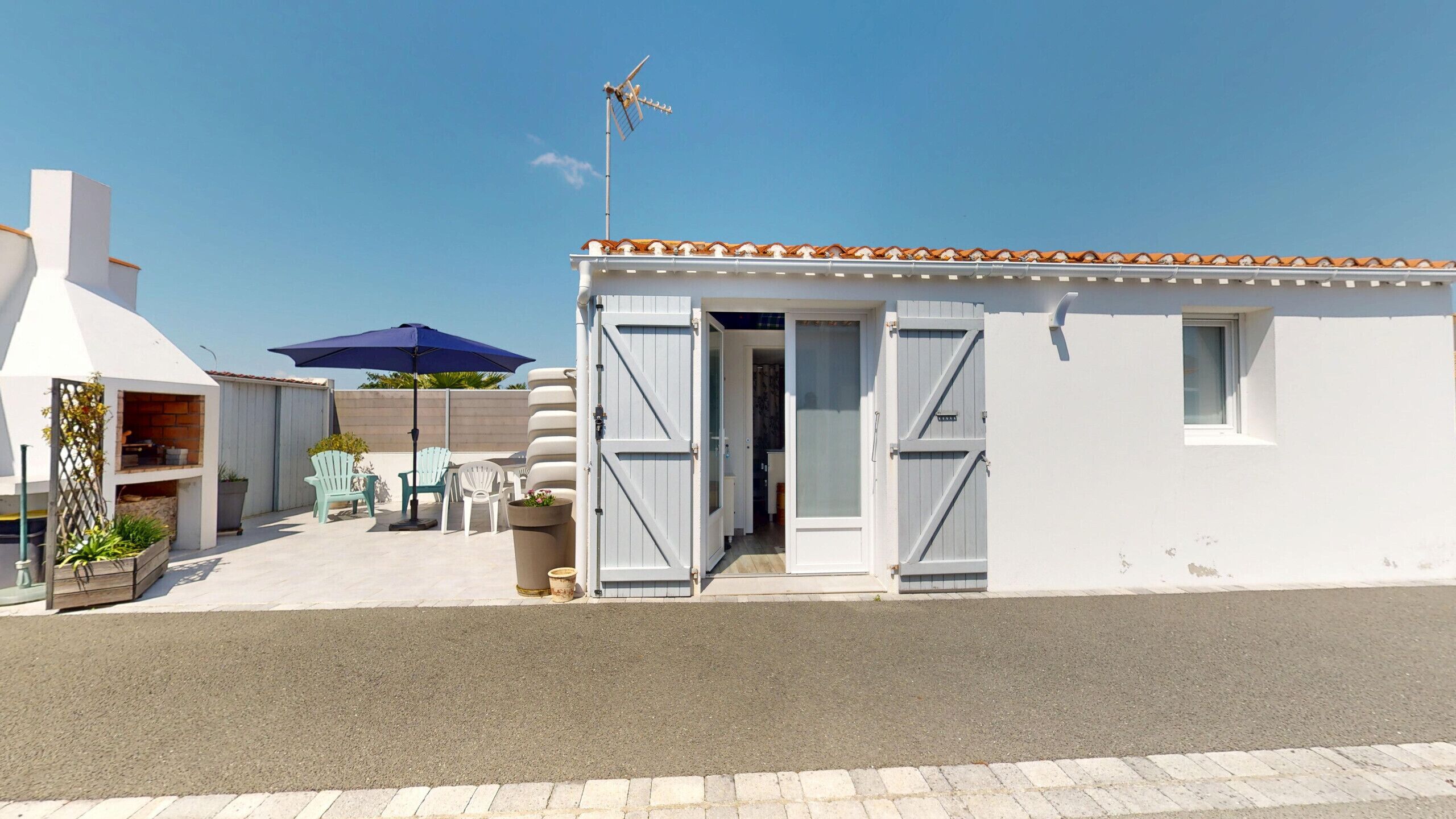 Een klein, wit gebouw met een pannendak en een grote open deur. Er staat een blauwe parasol en een terras met stoelen.