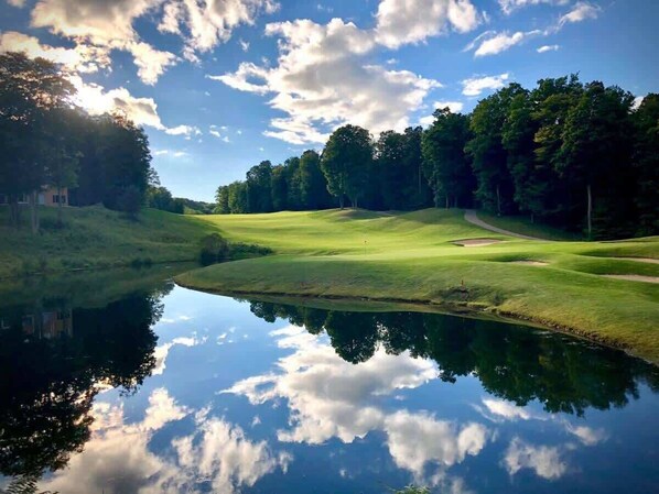 Ein friedlicher Golfplatz mit einem stillen See, der Himmel und Bäume spiegelt, umgeben von üppigem Grün.