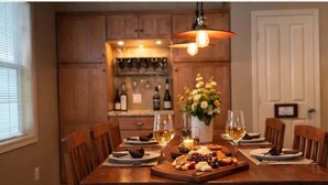 A dining table set with plates, glasses, and a cheese board, with wooden chairs and a hanging light fixture.
