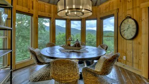 A wooden interior with a round table, wicker chairs, a chandelier, and a clock on the wall.