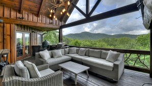 A covered outdoor seating area with a large sectional sofa, a small table, wicker chairs, and a view of a mountainous landscape.