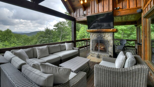 A wooden deck with a stone fireplace, wicker furniture, and a large window overlooking a forested landscape.