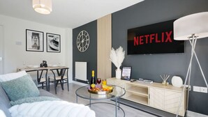 A modern living room with a sofa, a dining table, and a TV displaying the Netflix logo. - Welcoming House with Free Parking and Fast WiFi (Luton)