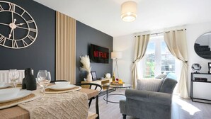 A modern living room with a dining area, a large wall clock, a Netflix sign, and a view of the city through large windows. - Welcoming House with Free Parking and Fast WiFi (Luton)