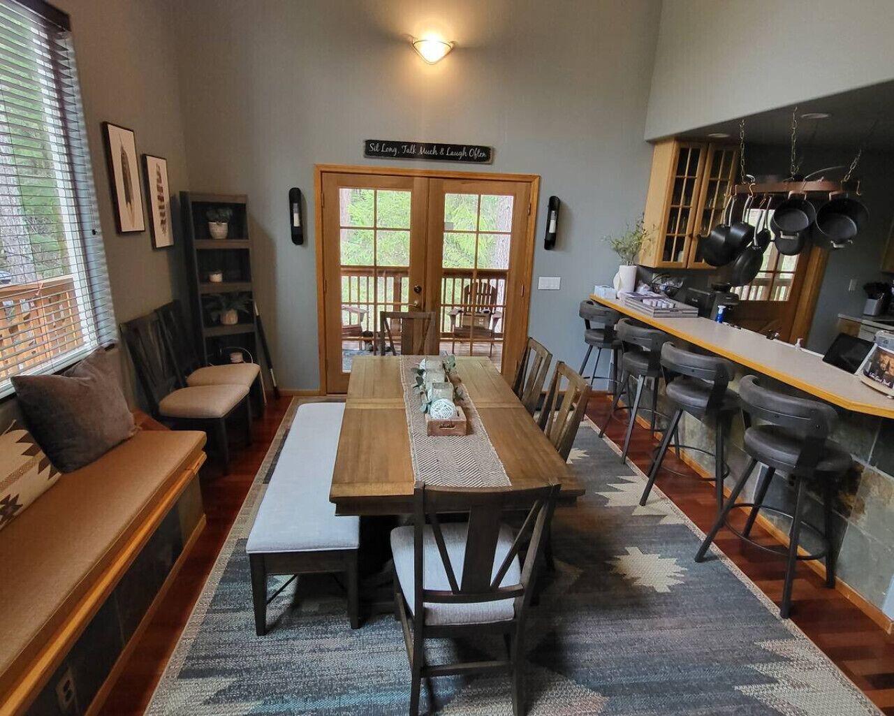 A dining area with a long wooden table, chairs, and a bench. There is a kitchen area with hanging pots and a sign above the door.